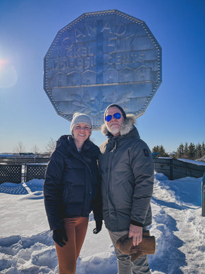 Two people standing in front of a large metal nickel on a snowy day.