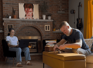 Two people in a cozy living room with a fireplace, one using a laptop and the other stringing an acoustic guitar.